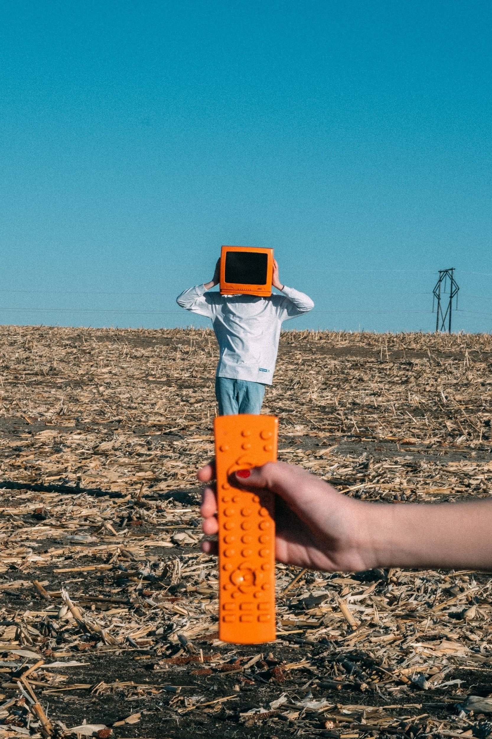 Photo of a person standing in a field of wooden debris with an orange television over their head, with a hand in the foreground holding an orange remote.