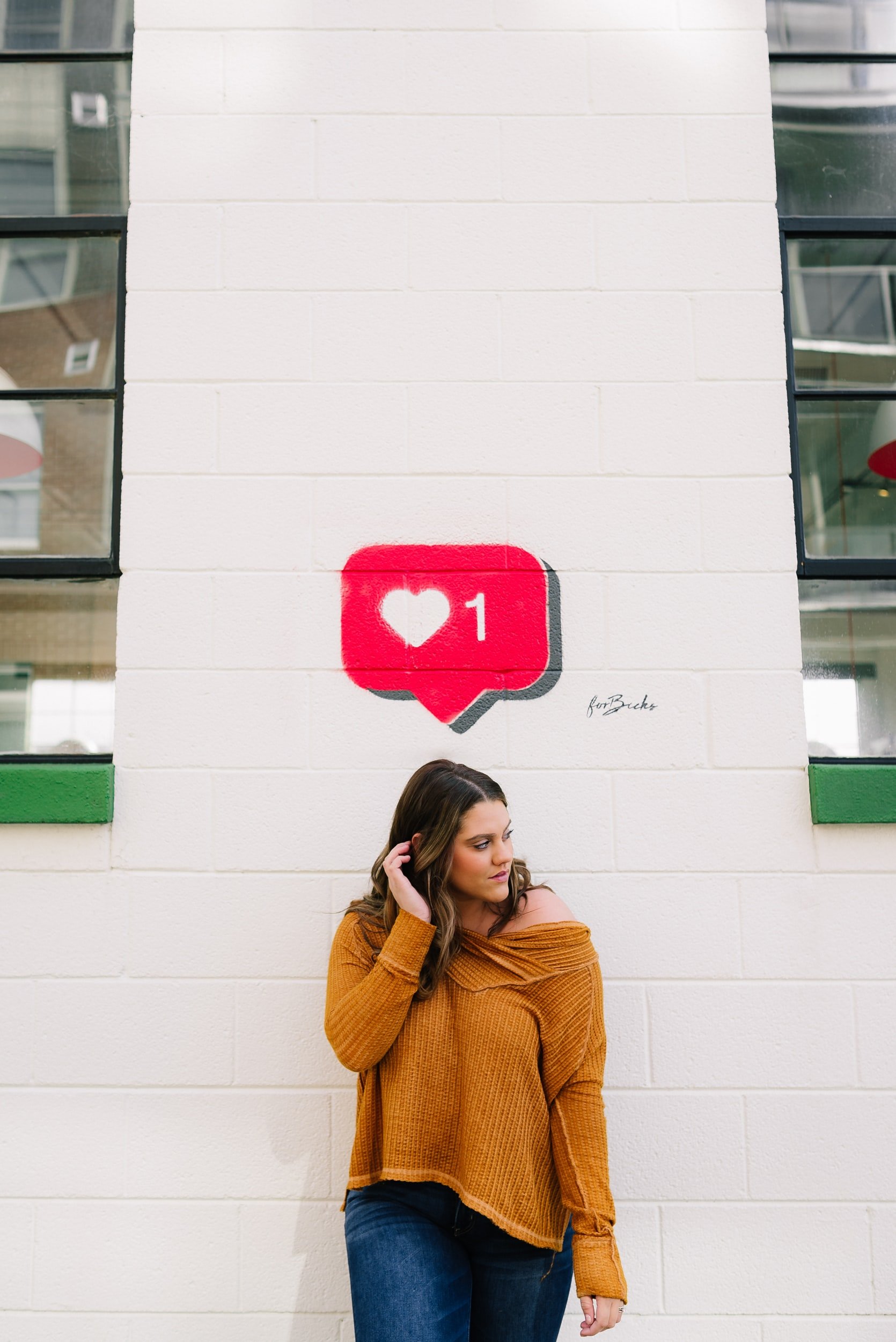 Photo of a young woman standing in front of a brick wall with a "Like" notification graphic painted on the section of wall above her head.